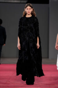 Female model walking on a red runway wearing a long black textured coat with feathered fabric detail on a dark backdrop