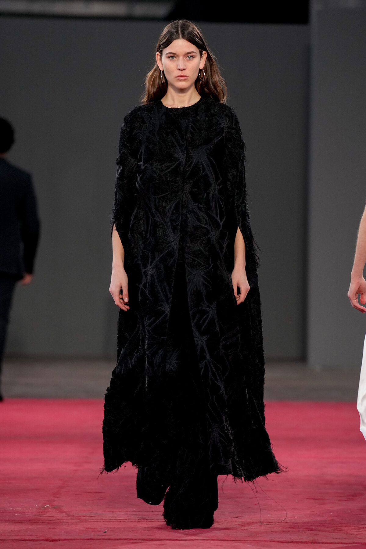 Female model walking on a red runway wearing a long black textured coat with feathered fabric detail on a dark backdrop