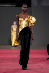Runway model wearing a gold metallic off-shoulder top with a large puff sleeve and black flowing pants on the catwalk.