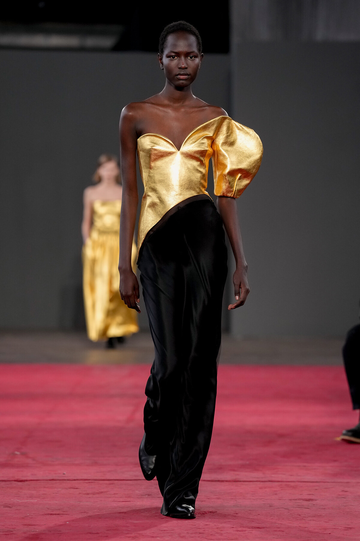 Runway model wearing a gold metallic off-shoulder top with a large puff sleeve and black flowing pants on the catwalk.