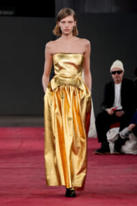 Model wearing a strapless metallic gold gown with a fitted bodice and sculpted peplum on a red runway.