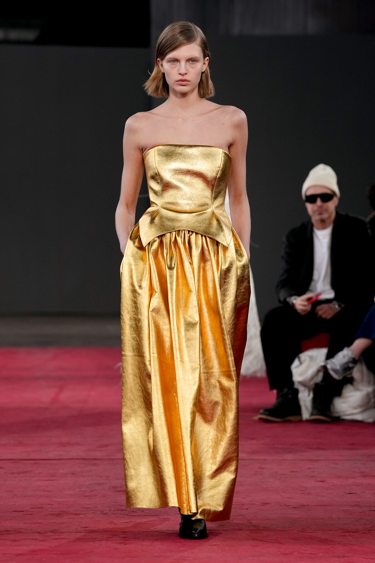 Model wearing a strapless metallic gold gown with a fitted bodice and sculpted peplum on a red runway.