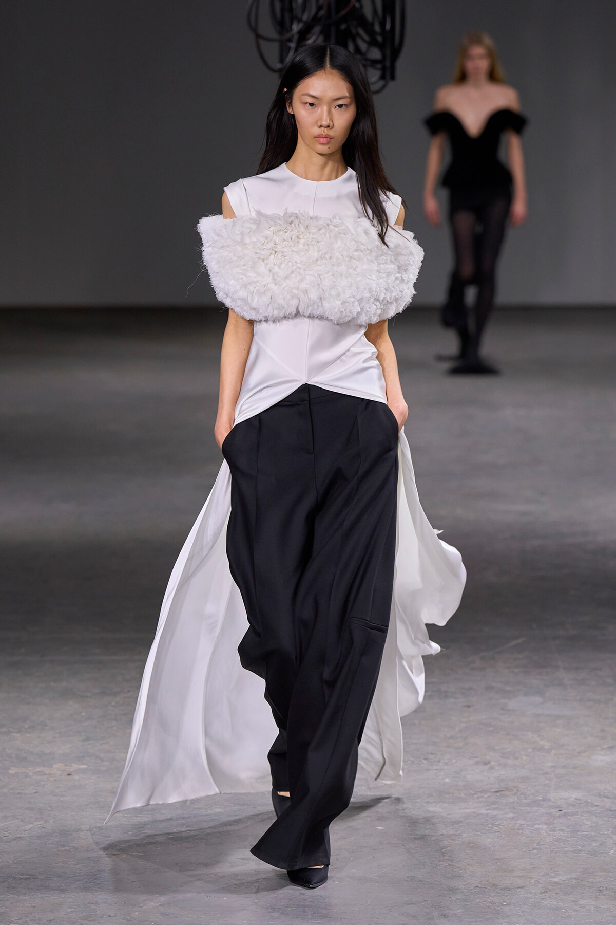 Model walks the runway in a white top with a large fluffy white shoulder piece and black wide-leg pants, cape trailing behind her.
