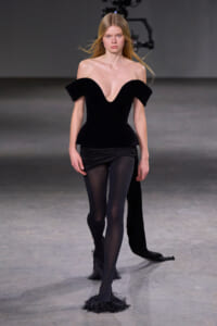 Runway model walking in a black off-the-shoulder peplum top, black tights, and furry black shoes on a gray runway.