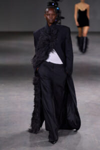 Model walking the runway in a black tailored suit with a long feathered scarf, white top peeking underneath, in an industrial setting with another model in the background.