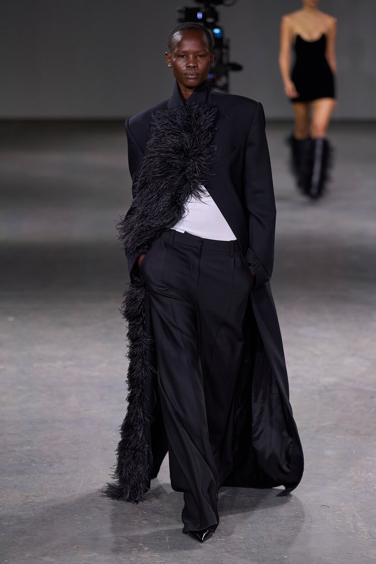 Model walking the runway in a black tailored suit with a long feathered scarf, white top peeking underneath, in an industrial setting with another model in the background.