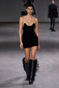 Runway model in a black mini dress with a nude illusion bodice and feather-trimmed knee-high boots.