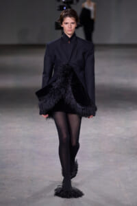 Model walking the runway in a black structured coat with feathered fringe at the hem and cuffs, against a neutral backdrop.