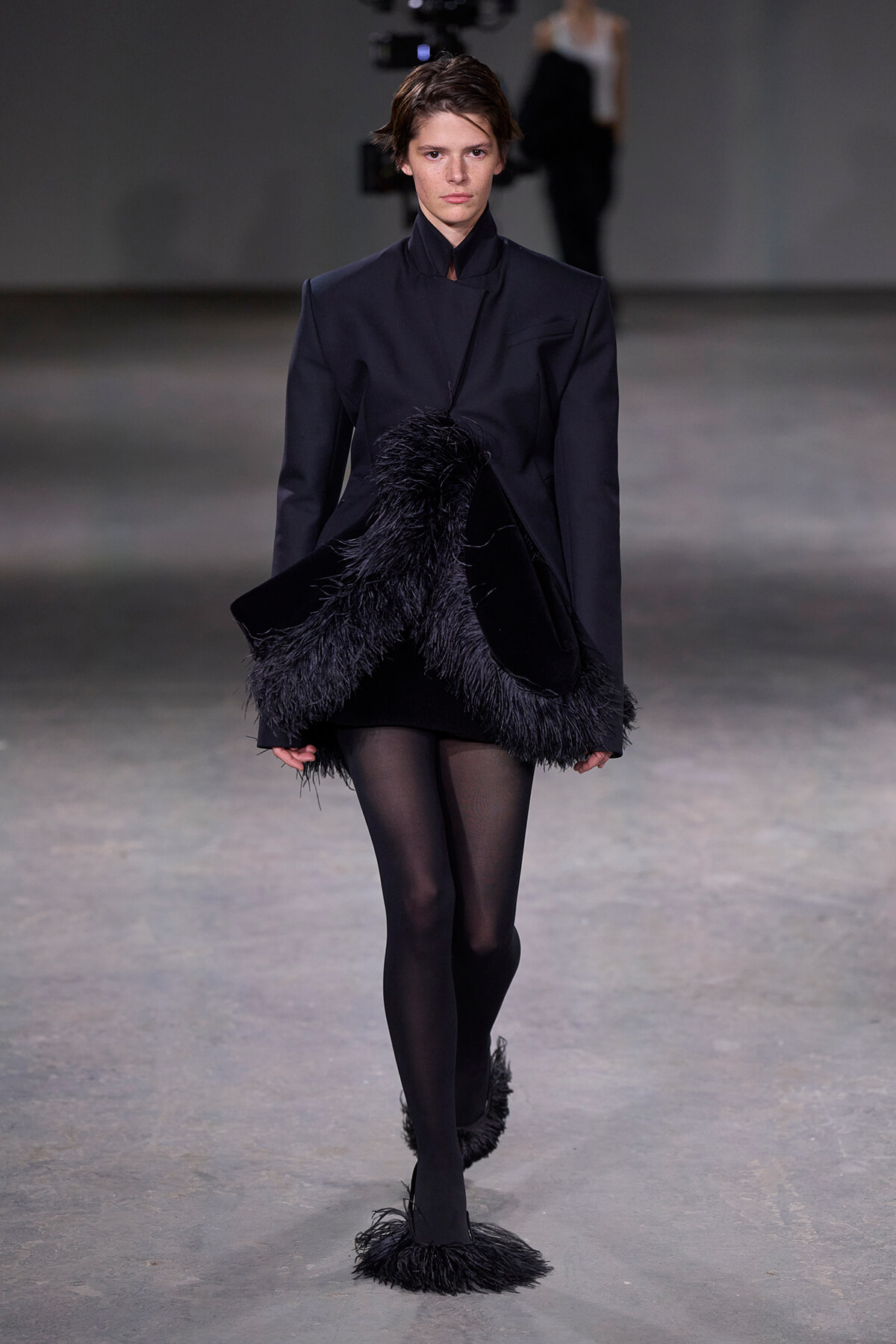 Model walking the runway in a black structured coat with feathered fringe at the hem and cuffs, against a neutral backdrop.