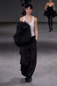 Model walks the runway in a white tank top and black wide-leg pants, holding a large black feathered garment over her arm.