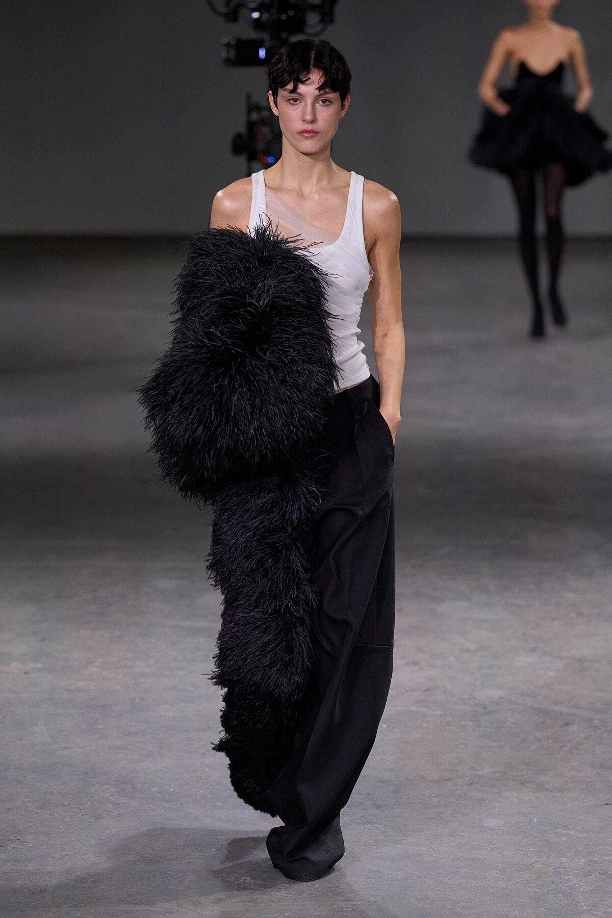 Model walks the runway in a white tank top and black wide-leg pants, holding a large black feathered garment over her arm.