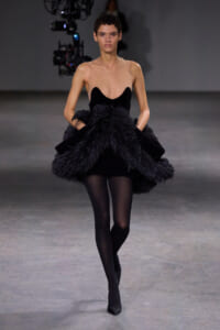 Female model walks a runway in a black sleeveless dress with a sheer nude bodice and a voluminous feathered skirt, paired with black tights and heels.