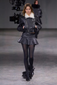 Model walking a runway in a black leather jacket with white shearling collar, matching leather mini skirt, sheer tights, and feathered boots.