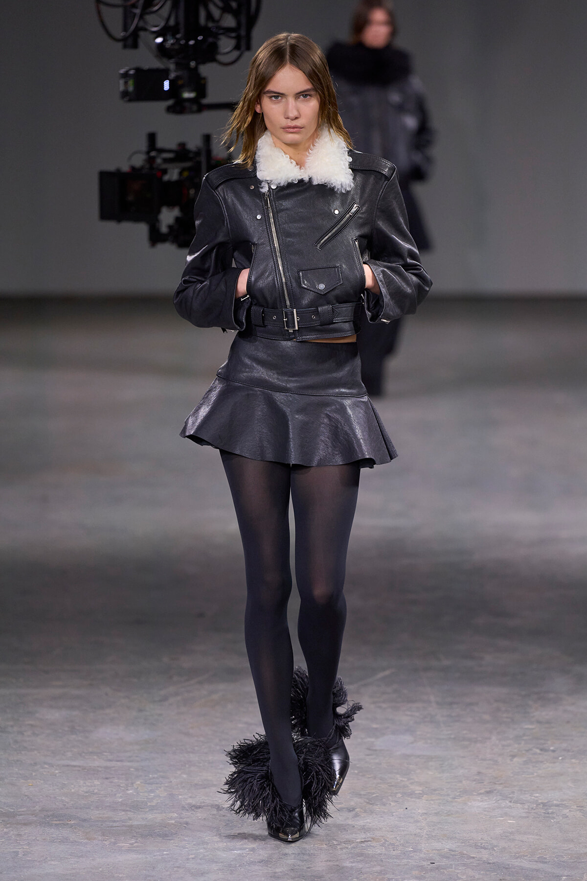 Model walking a runway in a black leather jacket with white shearling collar, matching leather mini skirt, sheer tights, and feathered boots.