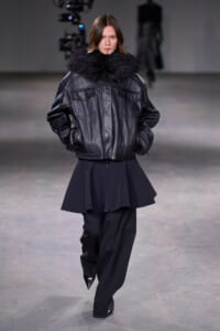 Model walks a runway in a black leather jacket with a thick fur collar, paired with black wide-leg pants and pointed boots.