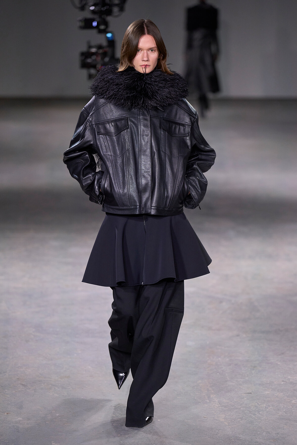 Model walks a runway in a black leather jacket with a thick fur collar, paired with black wide-leg pants and pointed boots.