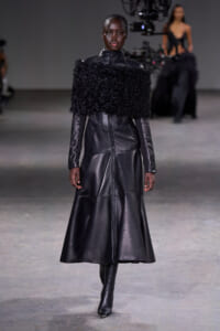 Female model wearing a long black leather coat with a fluffy fur collar on a runway,
