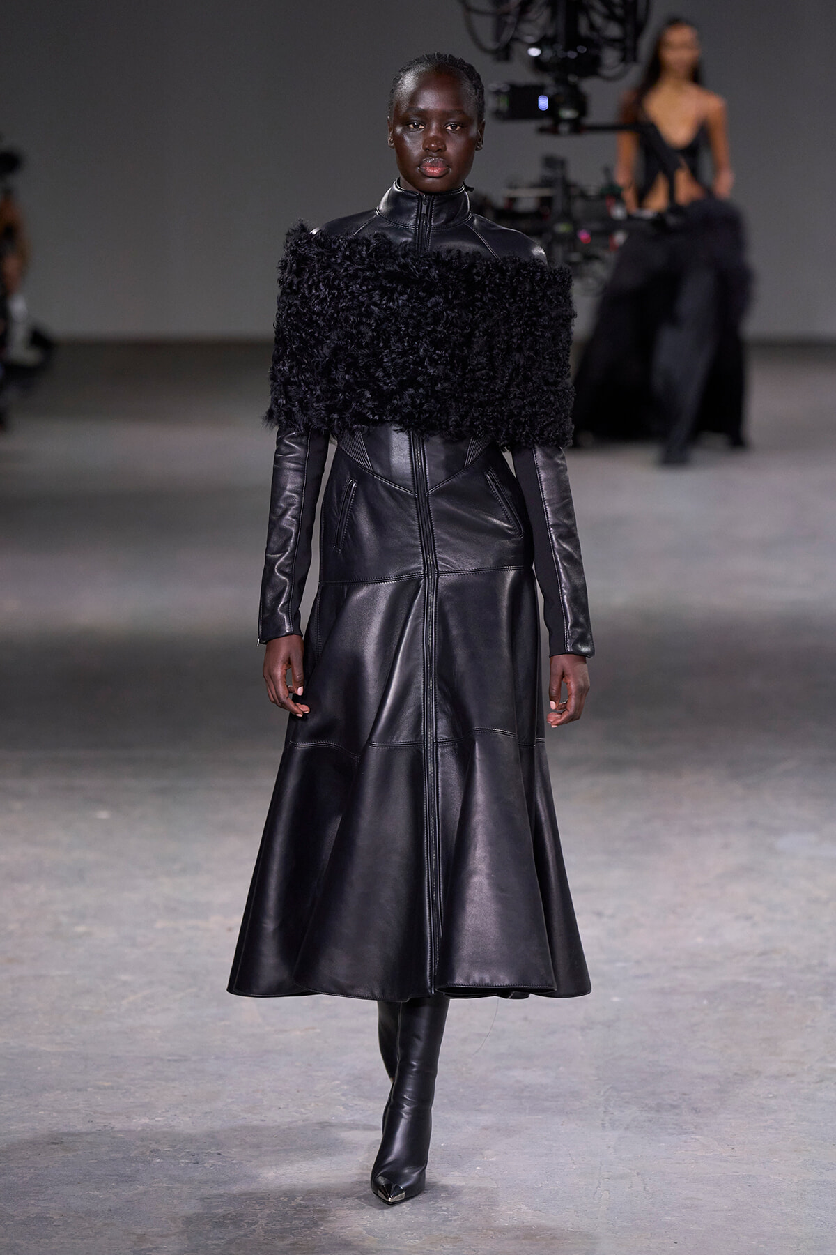 Female model wearing a long black leather coat with a fluffy fur collar on a runway,