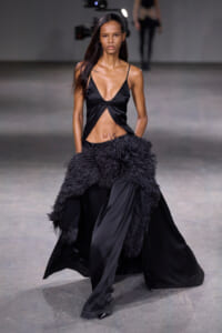 Model on a runway wearing a black satin outfit with a plunging V-neck and a large feathered wrap over the hips.