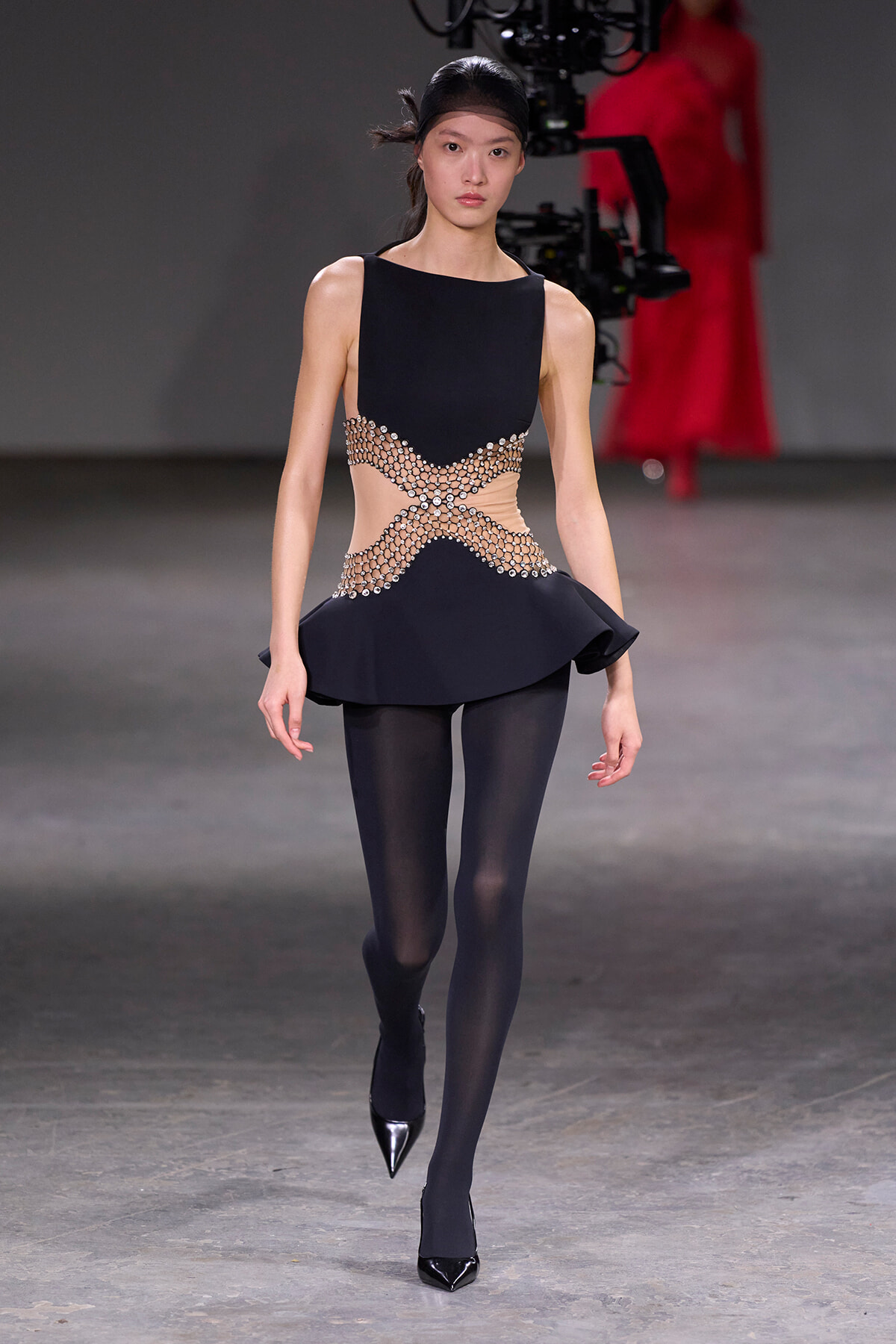 Model walking runway in a black dress with beige crystal lattice panels at the waist, black tights, and pointed heels.