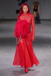 Model wearing a full-length red patterned gown with a large red feather accessory and red boots on a runway, carrying a silver chain bag.