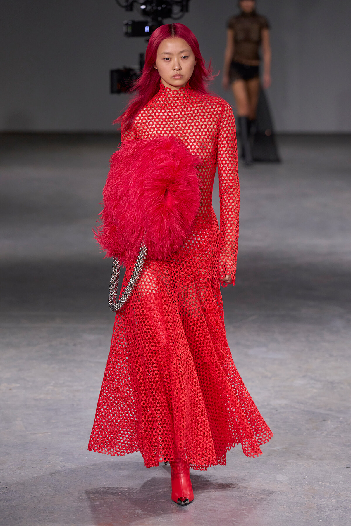 Model wearing a full-length red patterned gown with a large red feather accessory and red boots on a runway, carrying a silver chain bag.