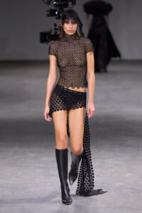 Model walking a runway in a black circular-cutout mesh top and matching shorts, pairing with knee-high black boots.
