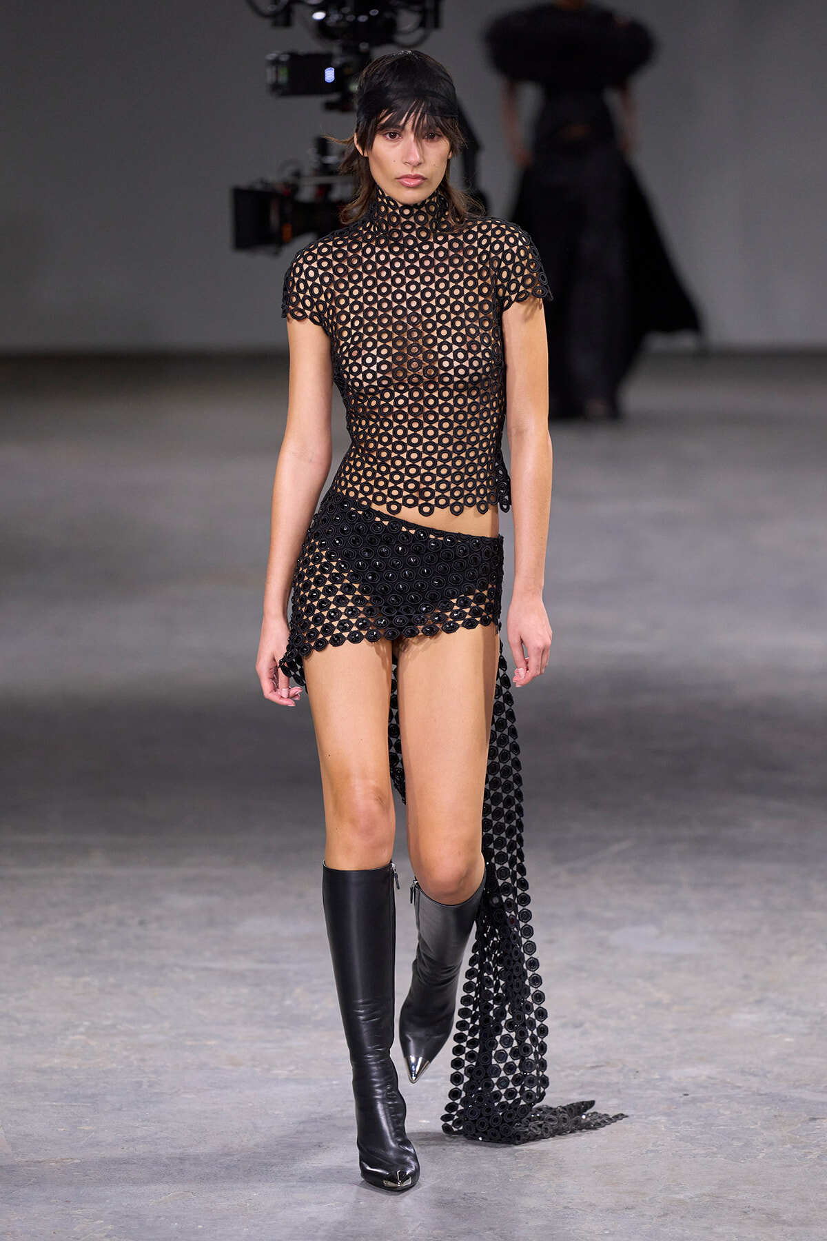 Model walking a runway in a black circular-cutout mesh top and matching shorts, pairing with knee-high black boots.