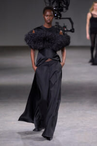 Model walks the runway in an all-black ensemble with a large black feather collar and wide-leg trousers, hands in pockets, on a dimly lit fashion show set.