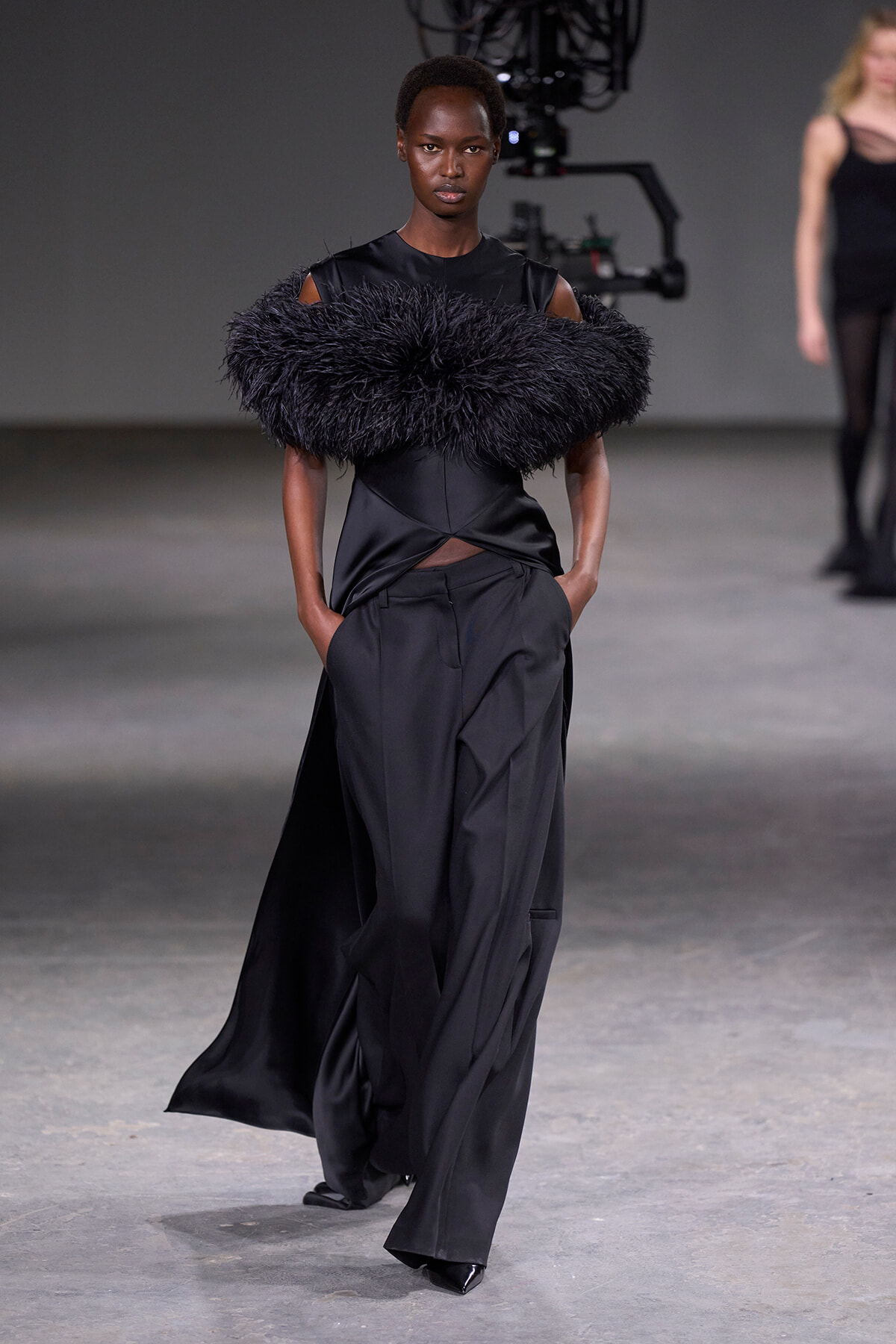 Model walks the runway in an all-black ensemble with a large black feather collar and wide-leg trousers, hands in pockets, on a dimly lit fashion show set.