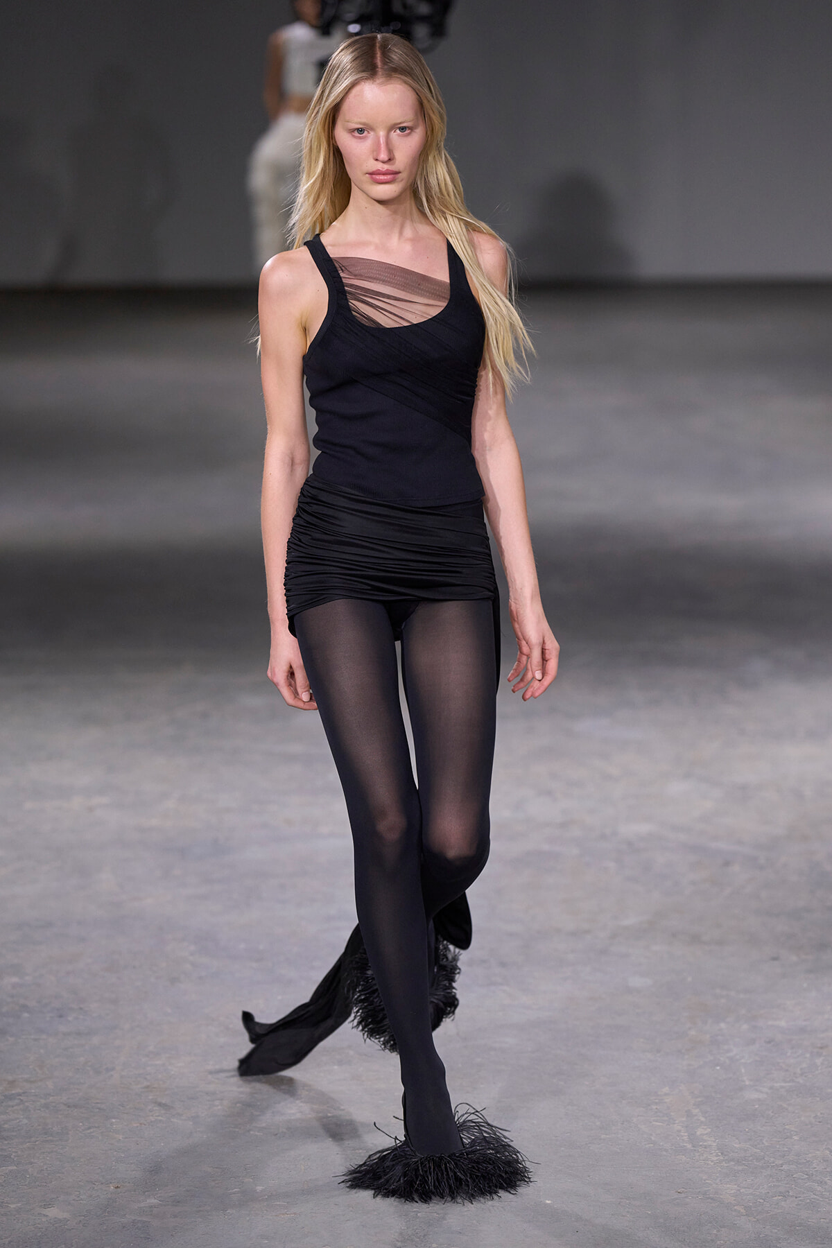 Model walks the runway in a black sleeveless top with a sheer mesh detail and a ruched black mini skirt, tights visible.