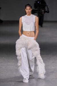 Model walks a fashion runway in a white sleeveless cropped top with a pleated fringe and matching white wide-leg pants, a fluffy white scarf draped around the hips.