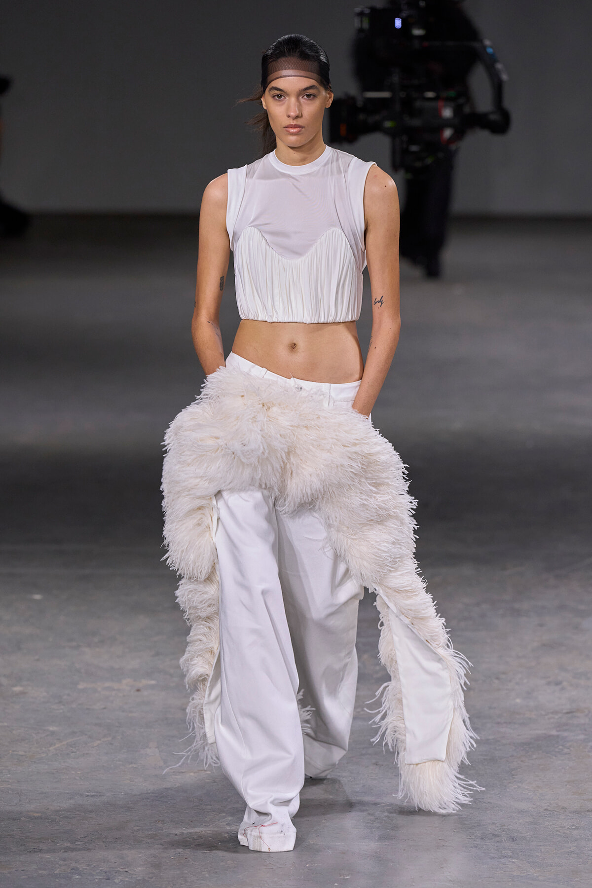 Model walks a fashion runway in a white sleeveless cropped top with a pleated fringe and matching white wide-leg pants, a fluffy white scarf draped around the hips.