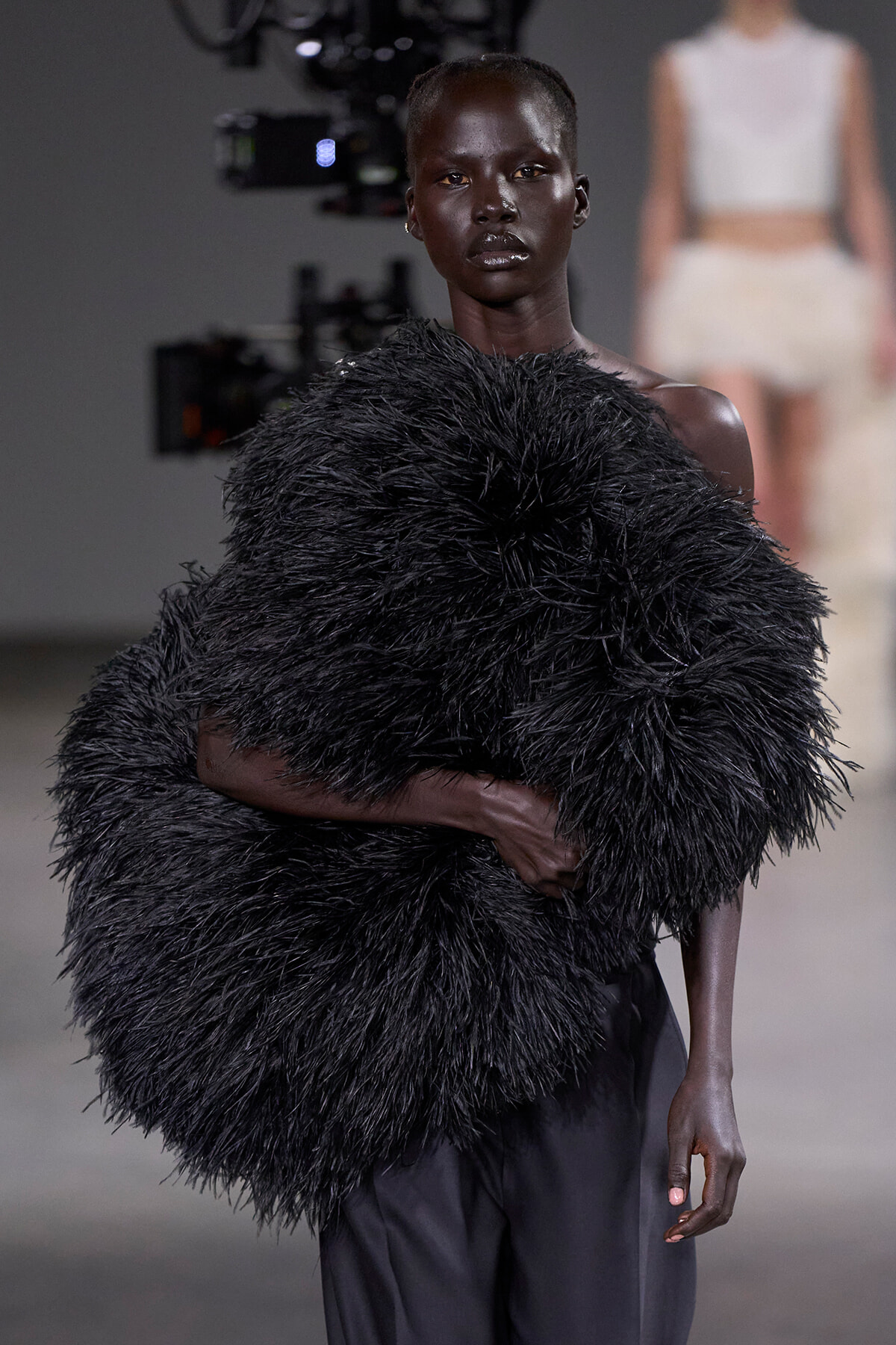 Model on a runway wearing a large black feathered shawl draped over the shoulders.