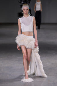 Model walks the runway in a white sleeveless top and a dramatic fluffy feathered skirt, with a sheer cap over her hair.