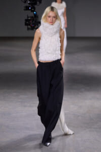 Model walks a fashion runway wearing a white fluffy turtleneck top and wide black trousers.