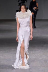 Model walking a fashion show in a white satin gown with a large fluffy feather collar and feathered hem, on a runway.