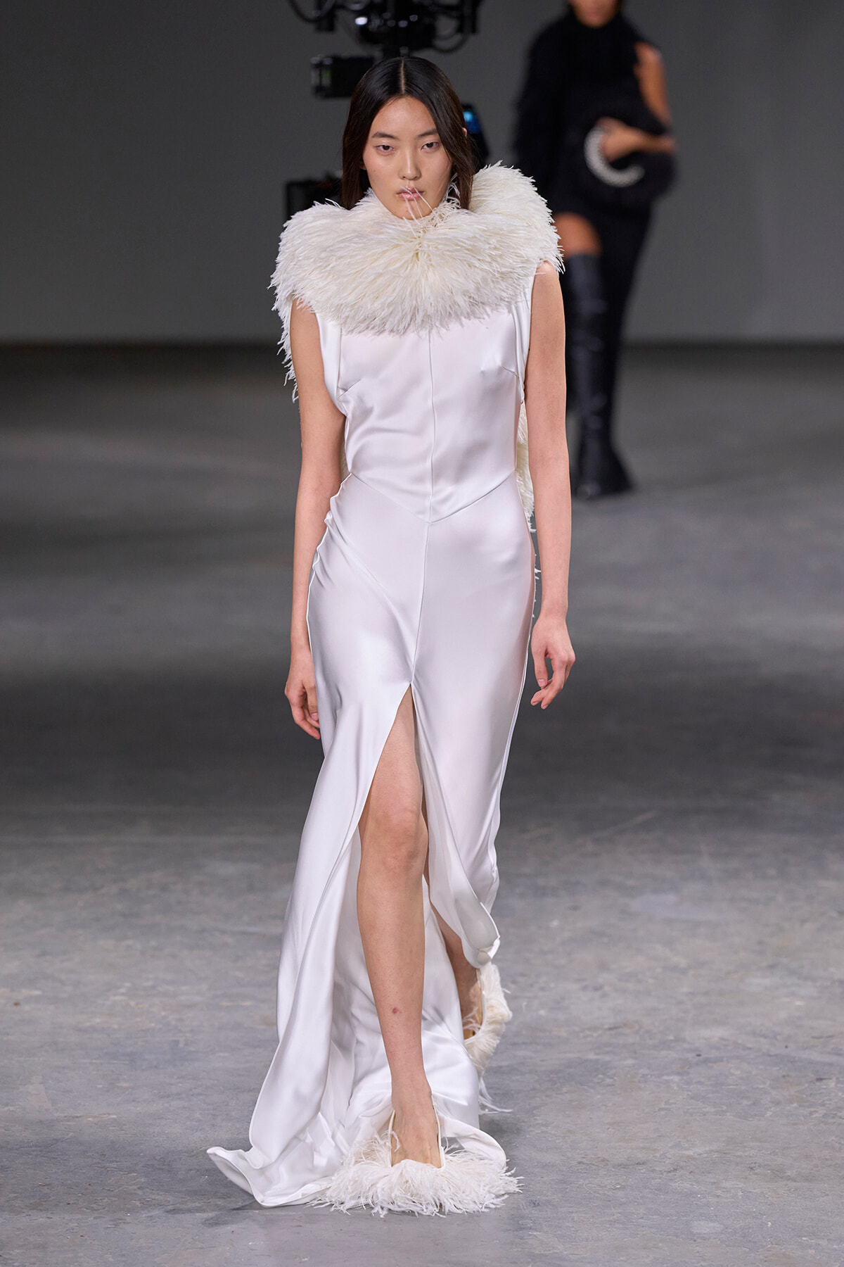 Model walking a fashion show in a white satin gown with a large fluffy feather collar and feathered hem, on a runway.