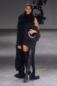 Model walks a runway in a black textured dress with a large shaggy black scarf and a circular fur clutch with silver trim.