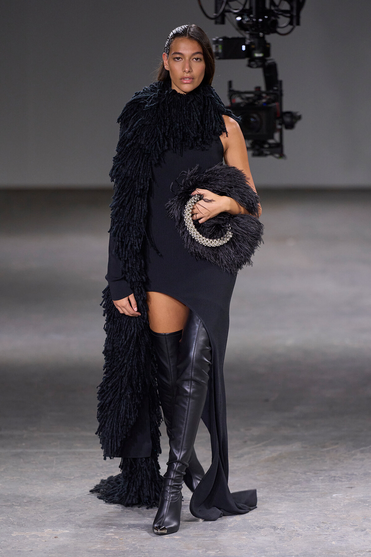 Model walks a runway in a black textured dress with a large shaggy black scarf and a circular fur clutch with silver trim.
