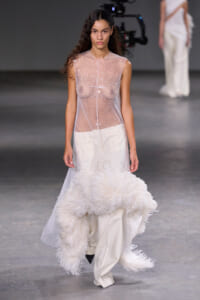Model walking the runway in a sheer, beaded sleeveless top over white pants, with a large white feathered accessory trailing behind her.