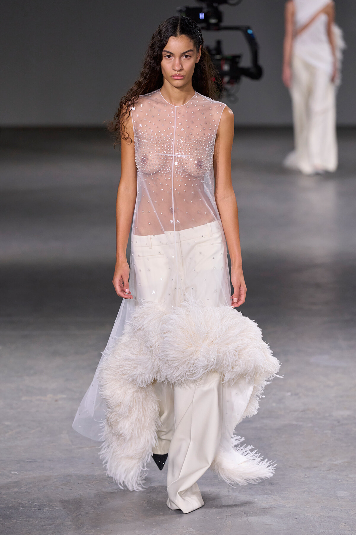 Model walking the runway in a sheer, beaded sleeveless top over white pants, with a large white feathered accessory trailing behind her.