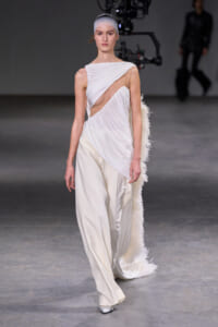 Model walks the runway in a white draped gown with a cutout bodice and feathered train.