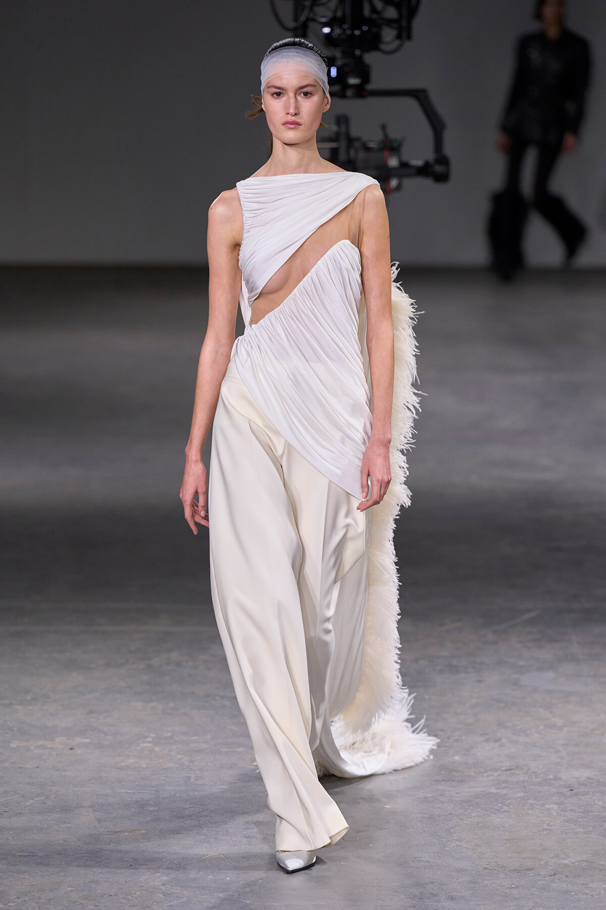 Model walks the runway in a white draped gown with a cutout bodice and feathered train.