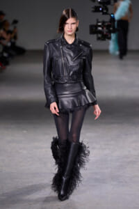 Model walks a fashion runway in a black leather biker jacket with a belt and a feathered mini skirt, paired with sheer tights and tall fur-trimmed boots.