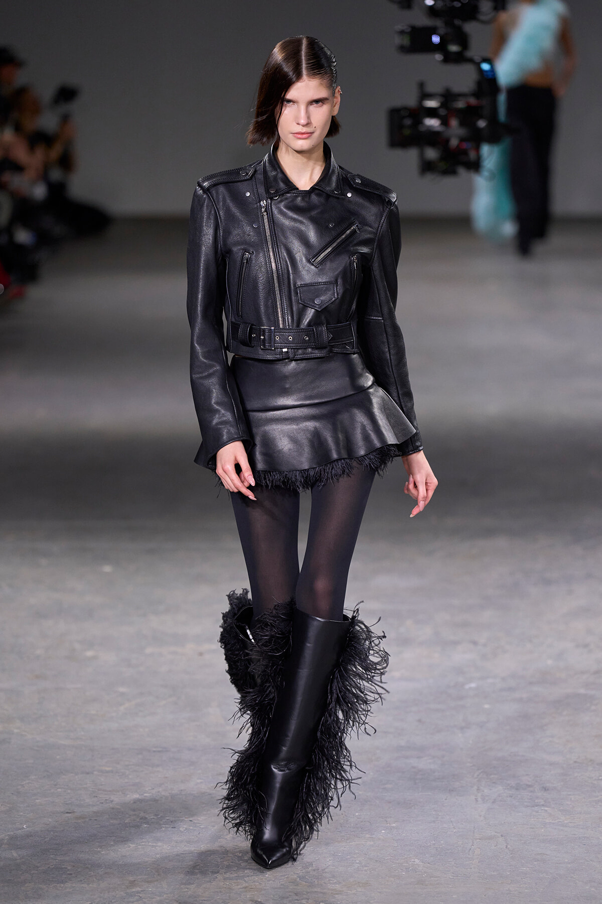 Model walks a fashion runway in a black leather biker jacket with a belt and a feathered mini skirt, paired with sheer tights and tall fur-trimmed boots.