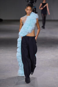 Model walking the runway in a light blue feather scarf draped over one shoulder with black tailored pants.
