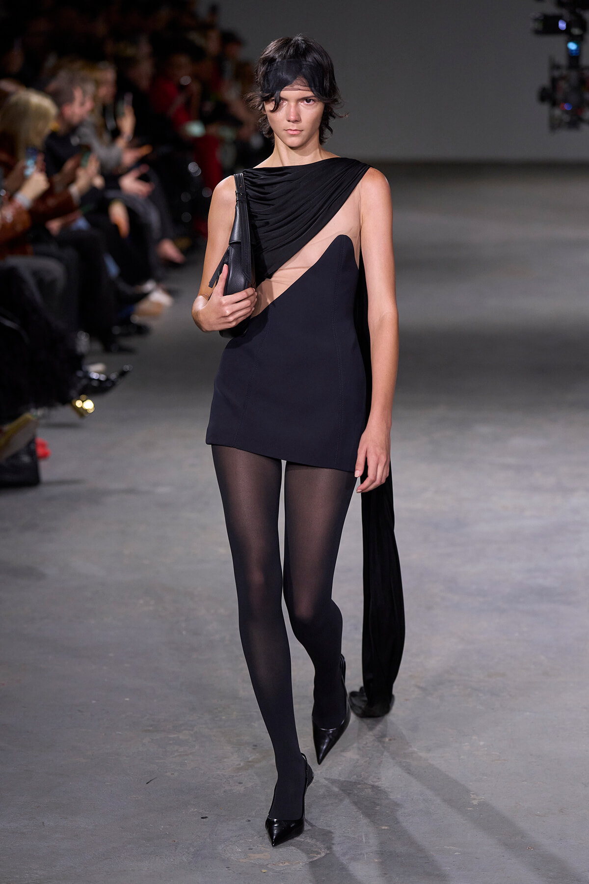 Model walks runway in a black asymmetrical mini dress with sheer panel and beige insert, tights and black heels, carrying a black bag
