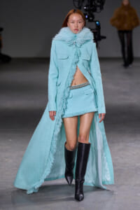 Model walks the runway in a pale turquoise textured coat with fringe edges, matching mini skirt, and knee-high black boots; scarf-like fur collar adds dramatic flair.