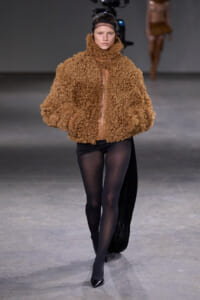 Model walking the runway in a tan, curly-textured coat with a center zipper, paired with black tights and pointed heels.